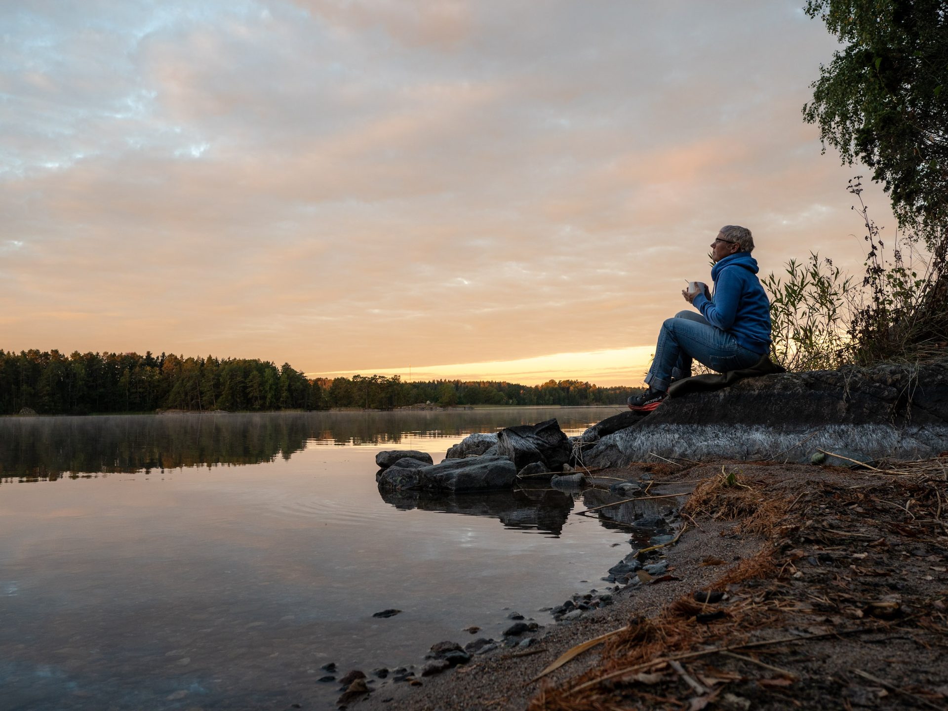 En person sitter på en sten vid sjön i gryningen med en kopp i händerna.