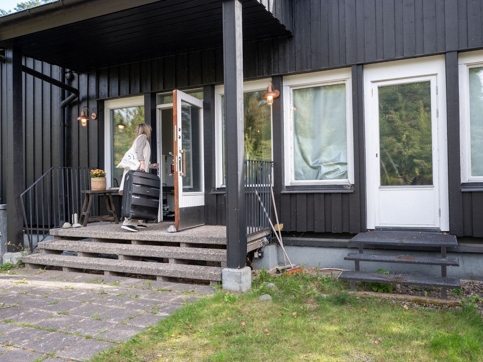 En person står i den öppna entrédörren till ett mörkt trähus.